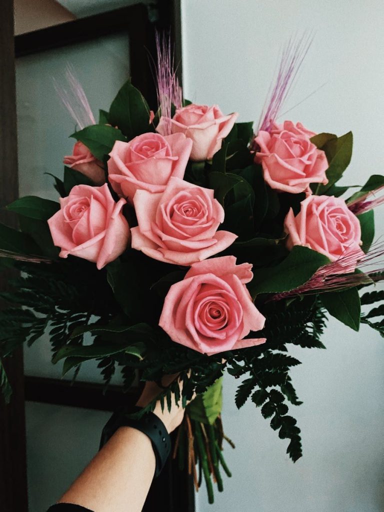 Crop anonymous person demonstrating bouquet of fresh pink roses with green branches tied with ribbon